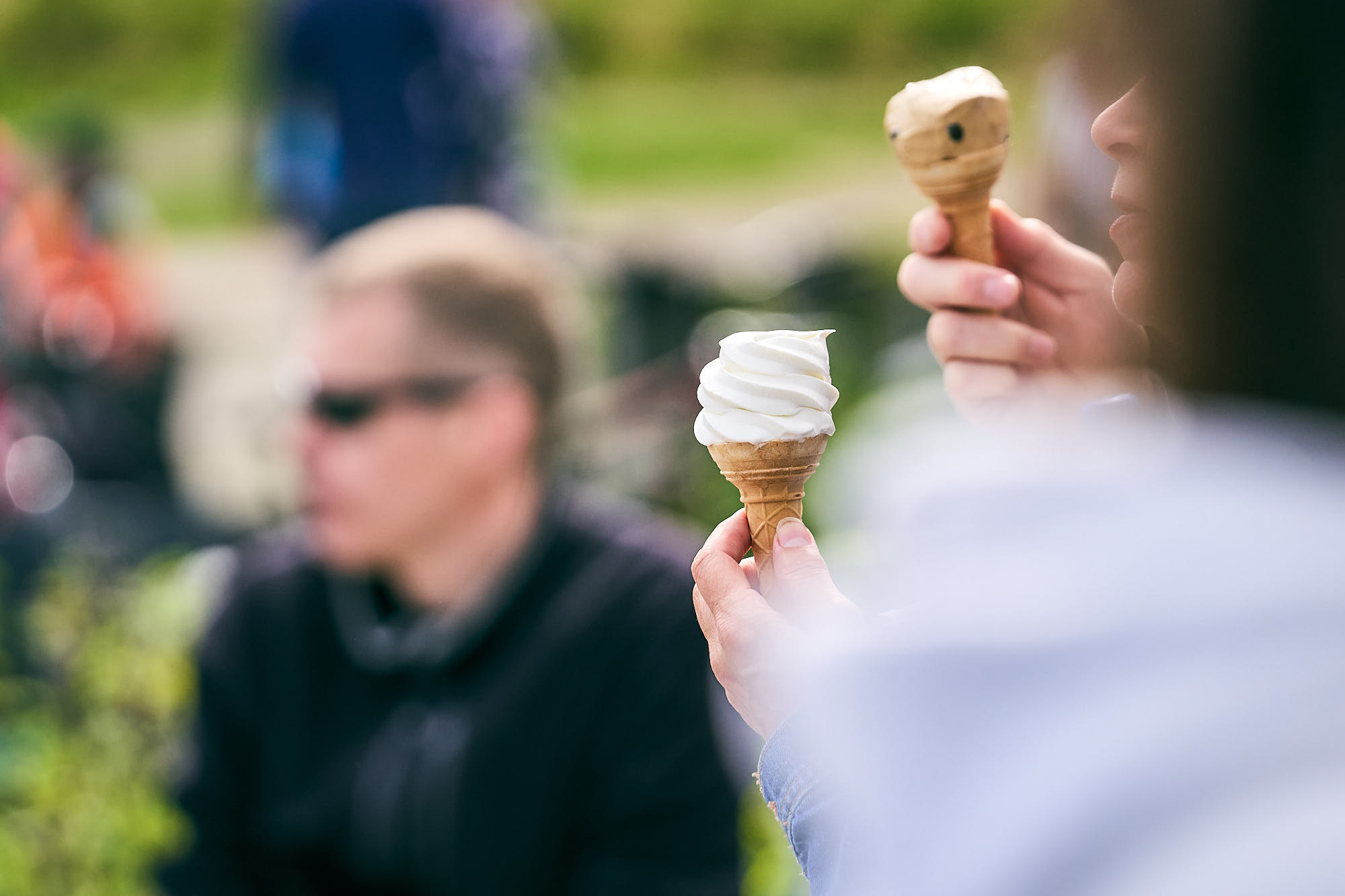 Ihmisen kädessä jäätelötötterö taustalla epätarkkoja ihmishahmoja ja kesäisen vihreä maisema.